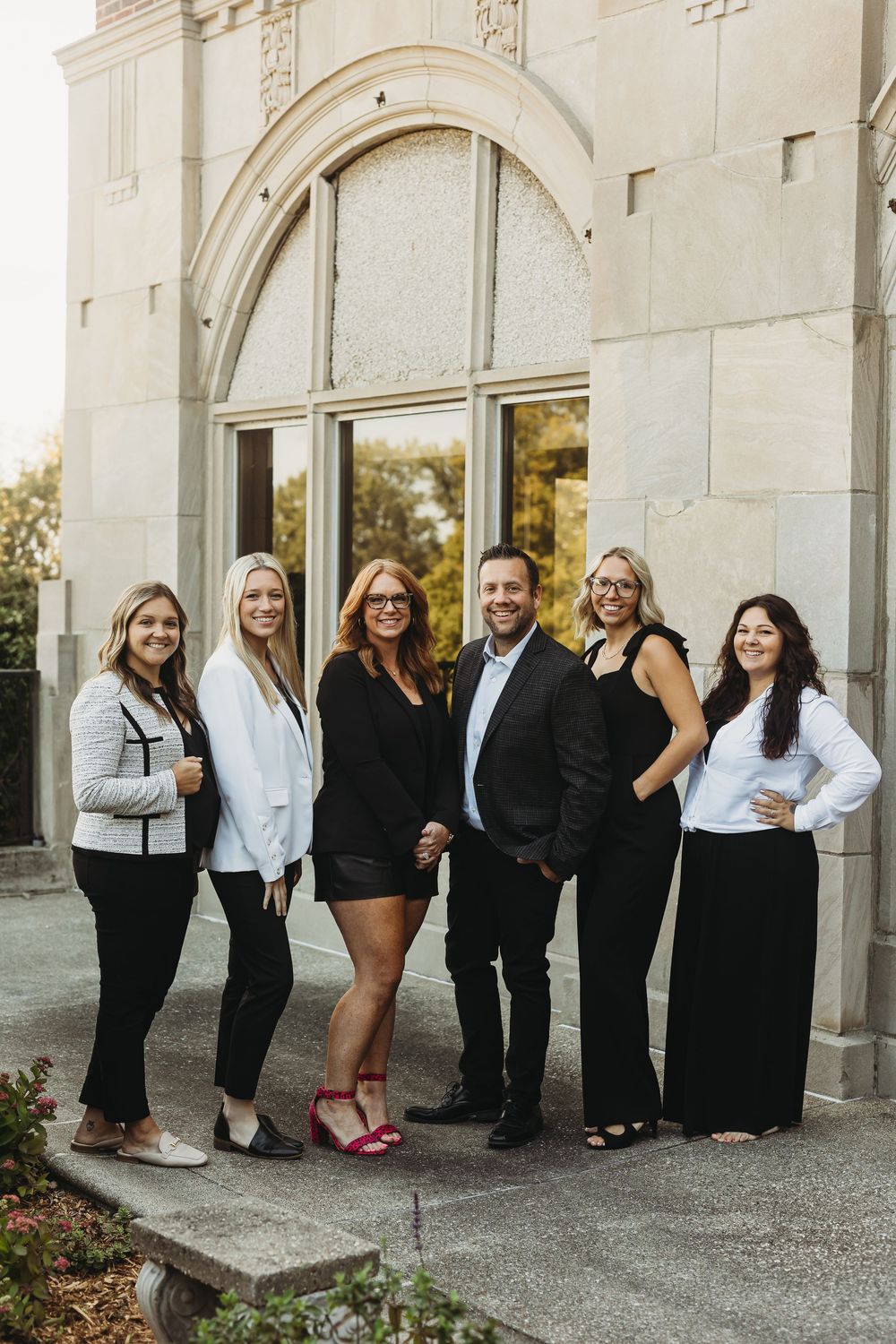 A group in coordinated black and white formal attire poses together outside a stone building with arched windows.