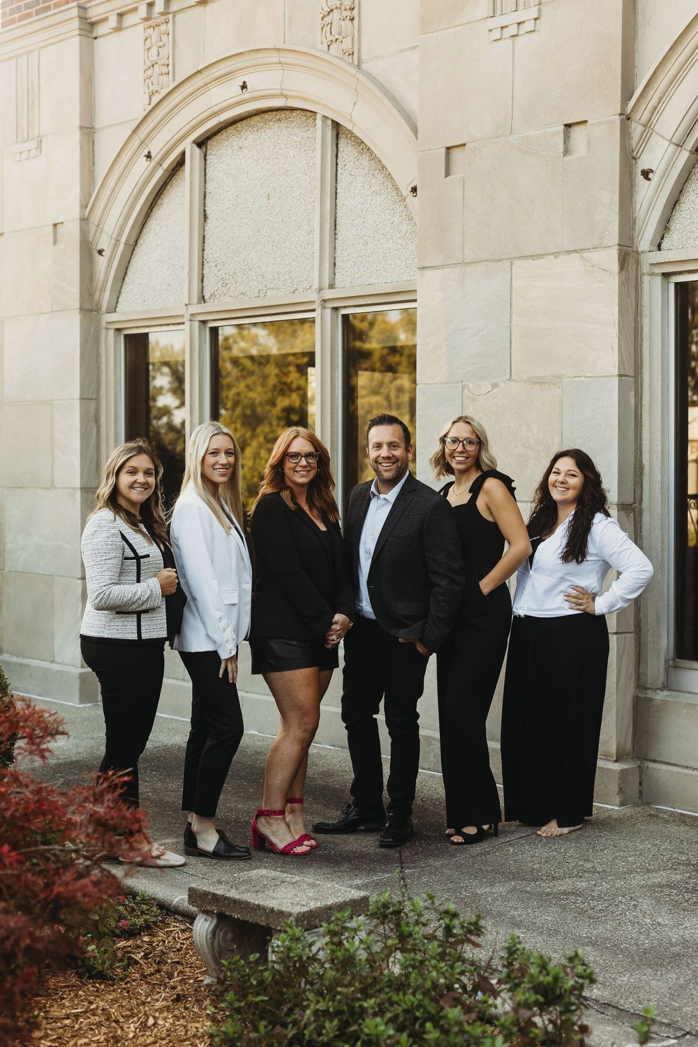 A group of people in formal black and white attire pose together against a stone building with arches and landscaping.