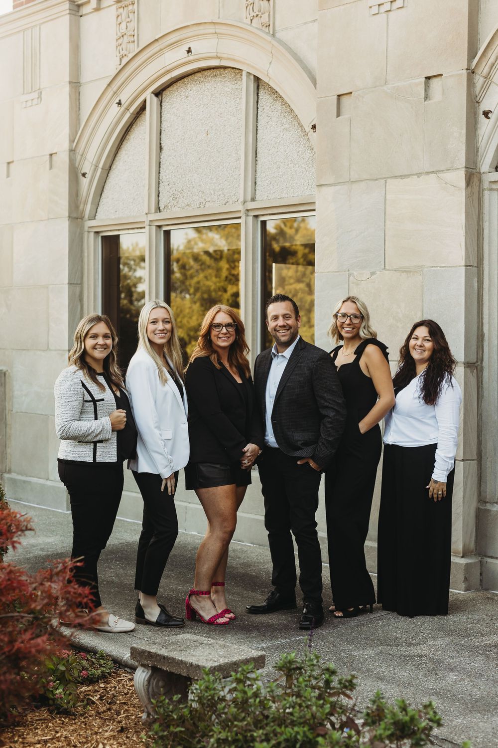 Wedding party members in black and white formal attire pose together against stone archways during autumn.