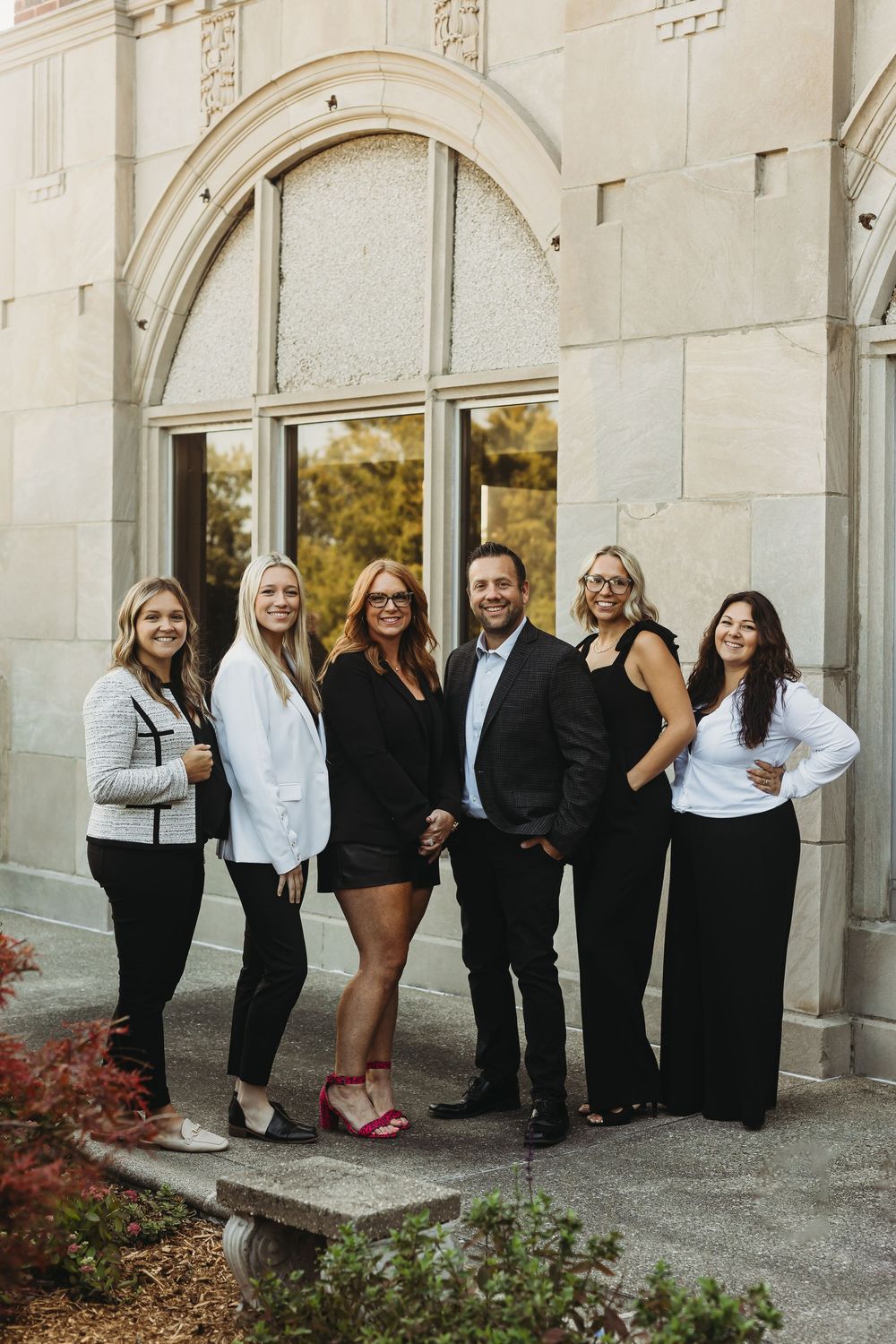 A group of people in formal black and white attire pose together in an outdoor archway setting.