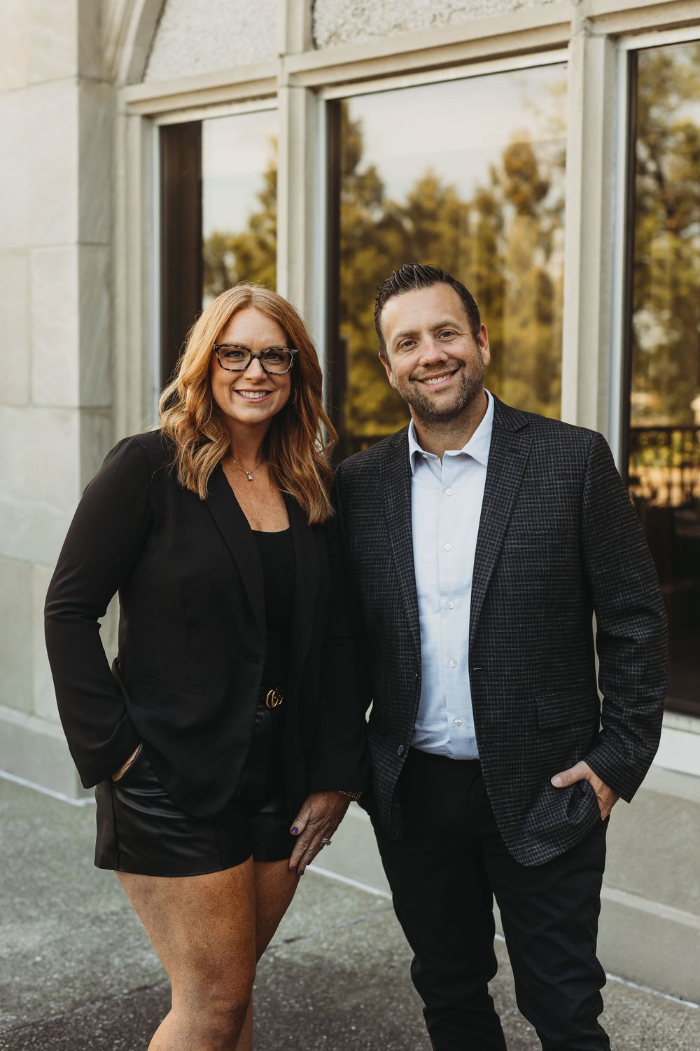 Professional couple in business attire poses together outside modern building in warm sunset lighting.
