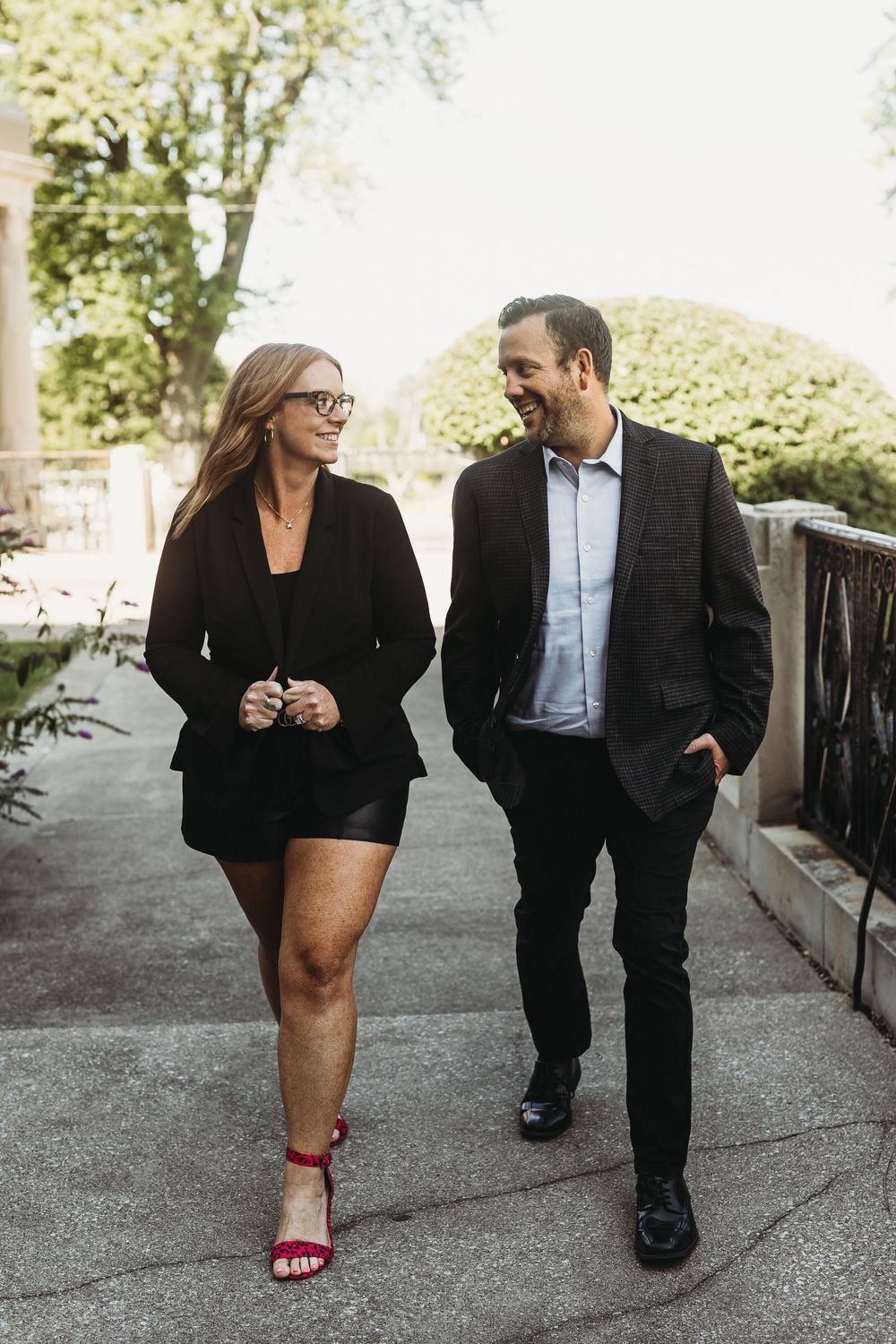 A couple in business attire walking and smiling together on a pathway surrounded by greenery.