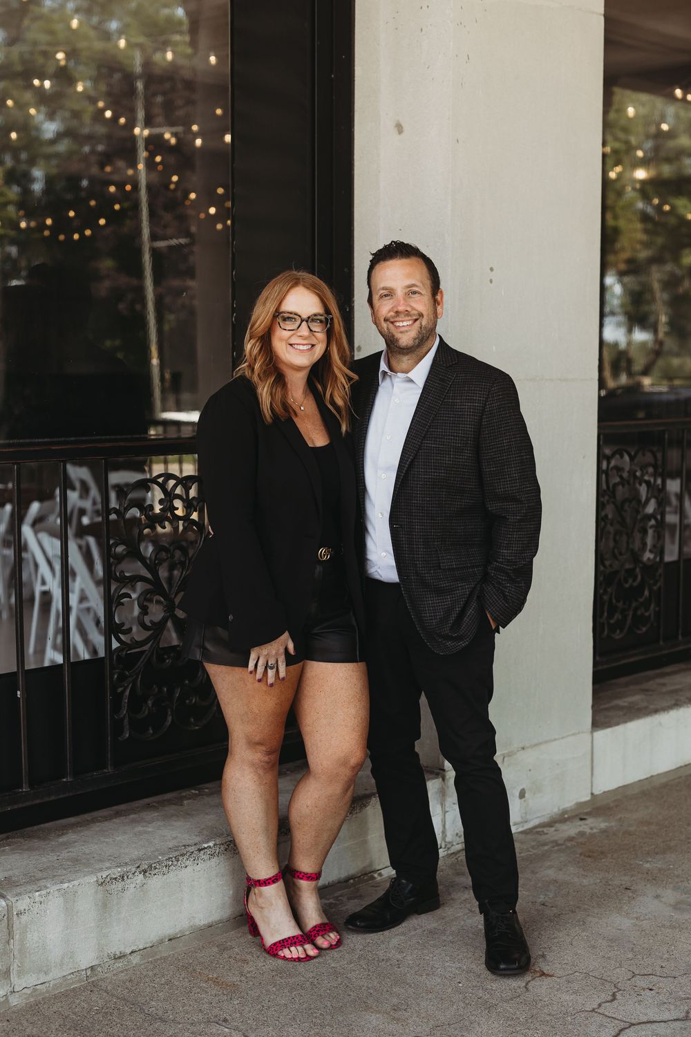 A couple poses together outdoors at dusk with twinkling lights visible in the background.