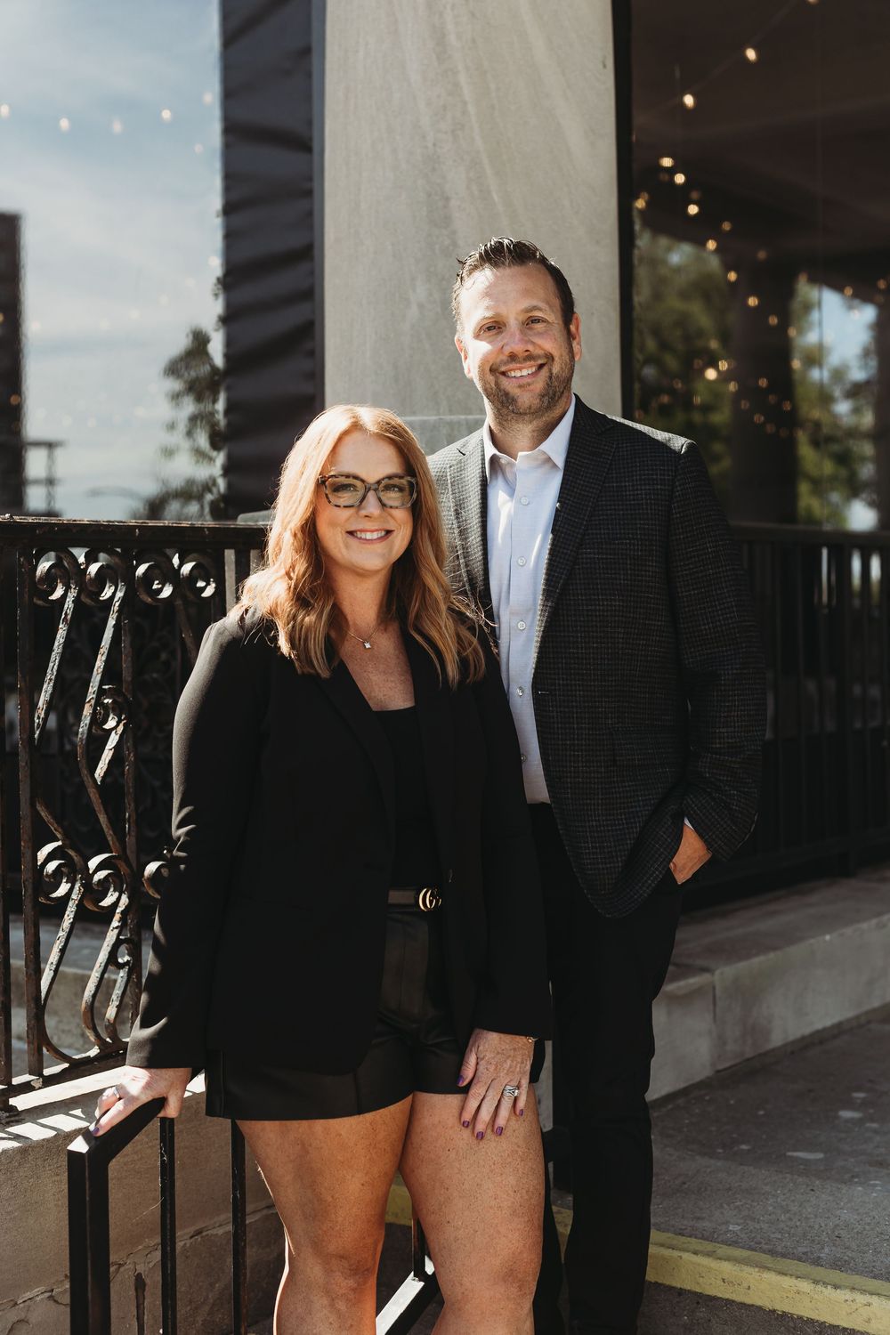 Couple poses together outdoors in formal attire with string lights and architectural elements in background.