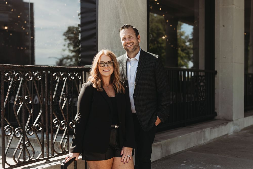 A couple stands together on an elegant porch with ornate black railings during an outdoor photoshoot.
