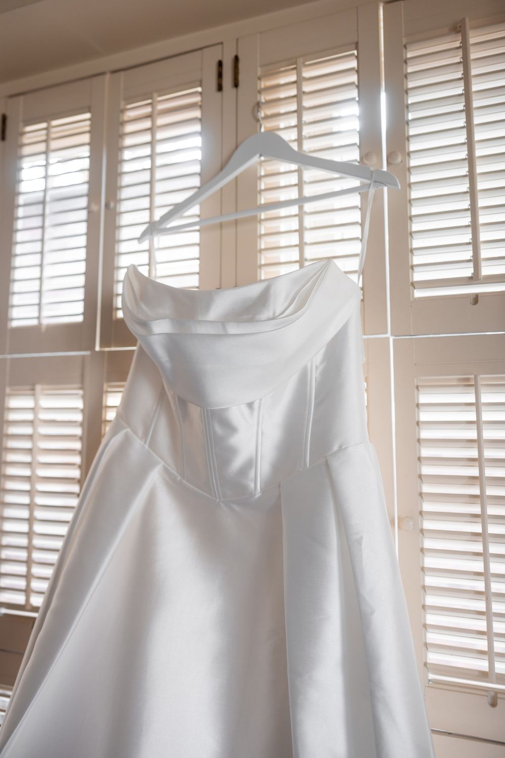 Row of white bridesmaid dresses hanging on hangers against plantation shutters in natural light.
