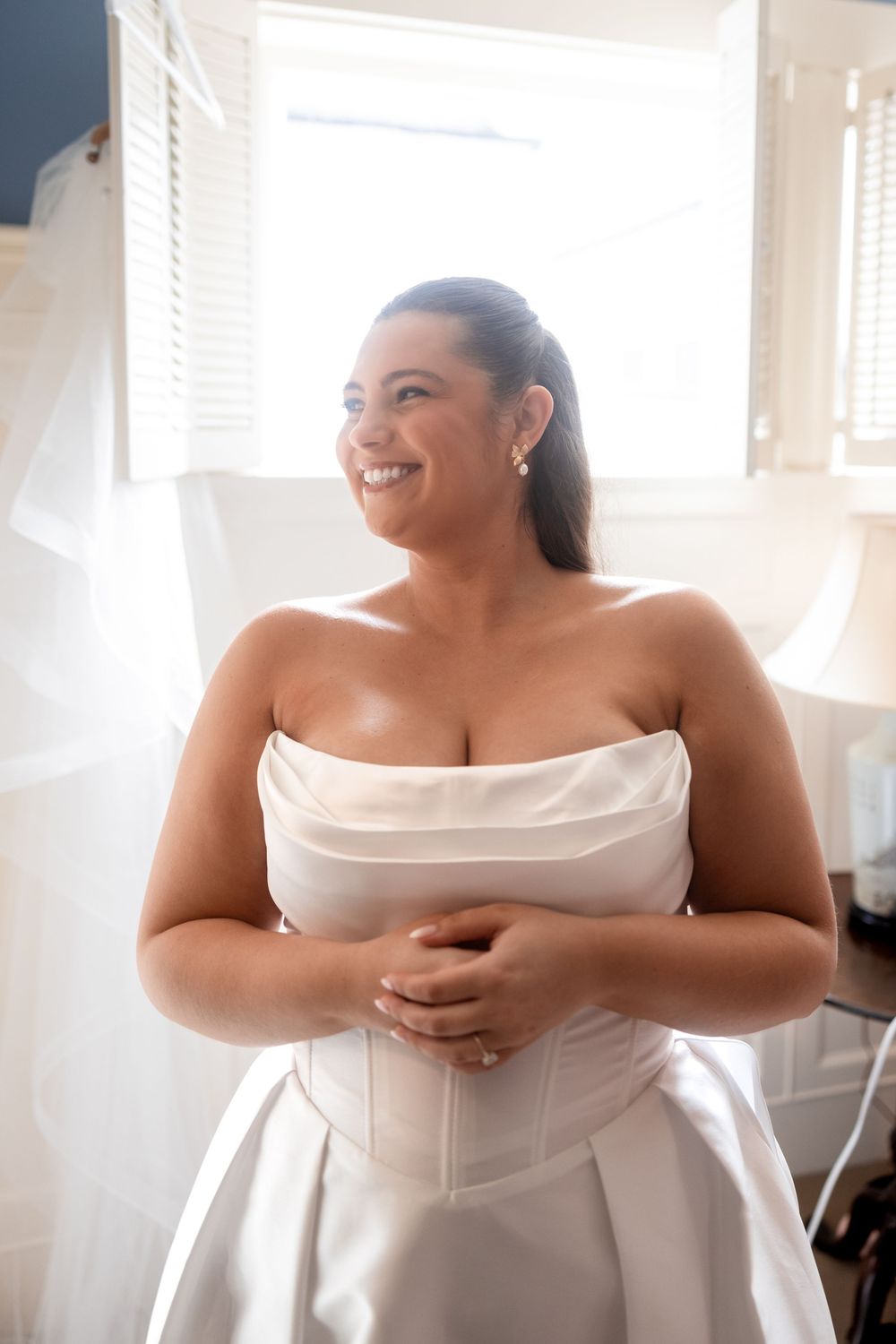 Bride in white strapless dress smiling joyfully while getting ready in sunlit room.
