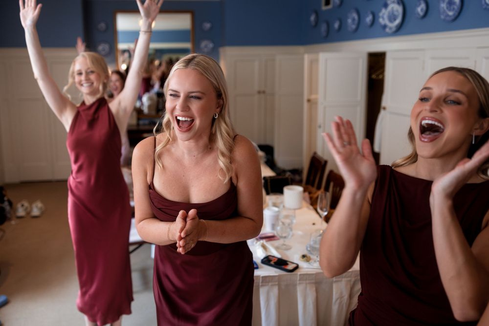 Bridesmaids in burgundy dresses celebrate with raised arms and joyful expressions.