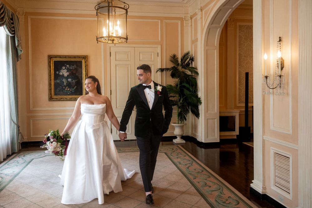 A couple in wedding attire walks hand in hand through an elegant hallway with ornate lighting and peach-colored walls.