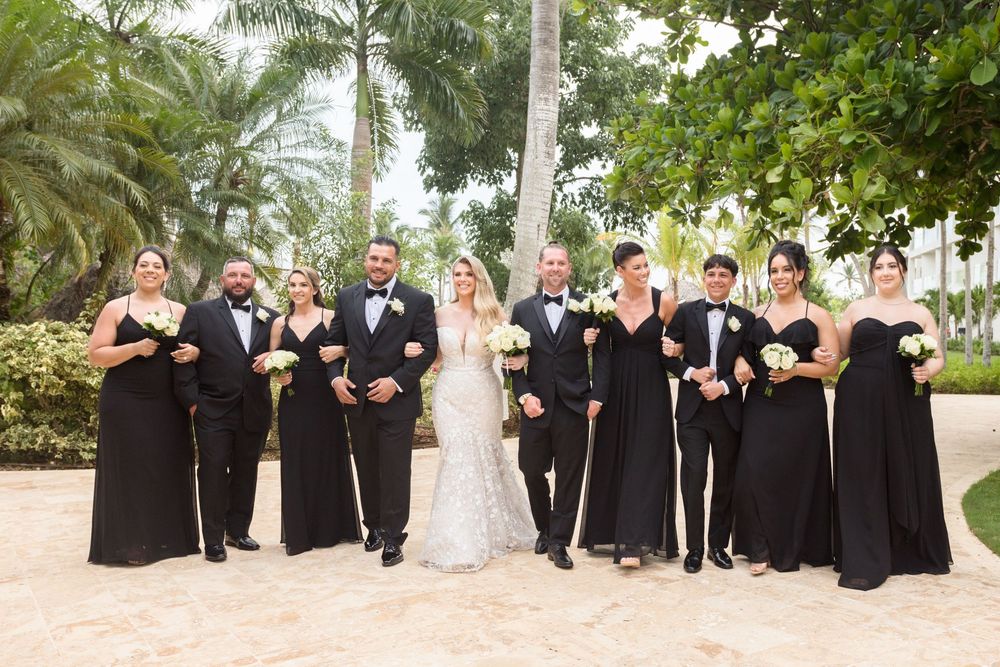 Wedding party in black and white attire poses on grass with palm trees in the background.