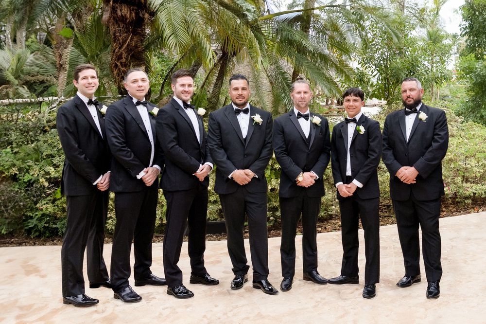 A group of seven men in black tuxedos and bow ties stand together in a garden setting with palm trees.