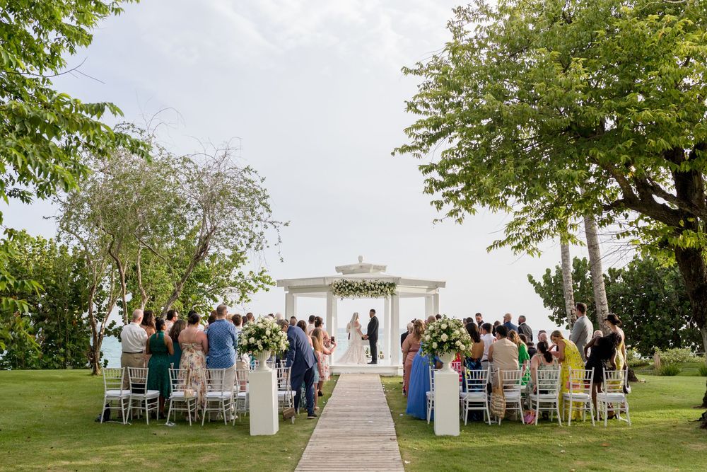Outdoor wedding ceremony setup with white chairs and gazebo surrounded by lush green trees.