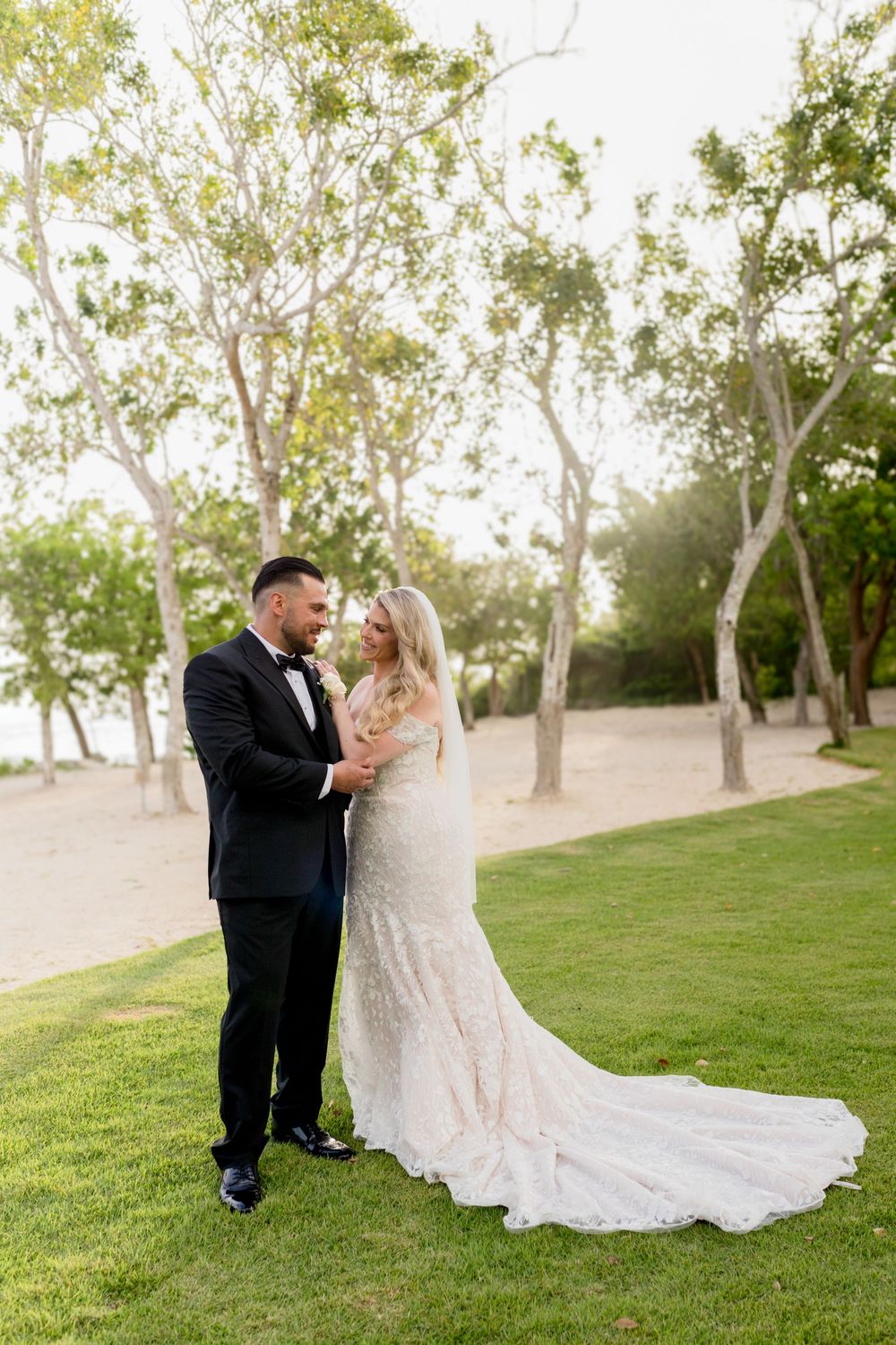 A couple shares a romantic moment in their wedding attire on a grassy lawn with tall trees in the background.