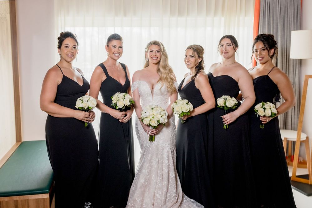 A wedding party wearing black dresses and white gowns poses together in a hotel room for formal photos.