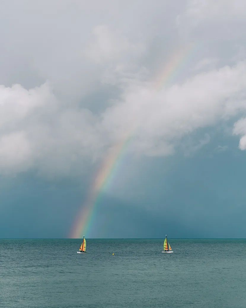 Rainbow forming over the calm sea with two yellow sailing boats beneath it.