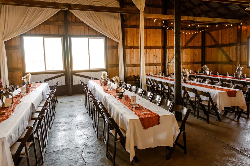 Long rows of elegant tables and chairs set up in a rustic barn venue with white linens and string lights overhead.