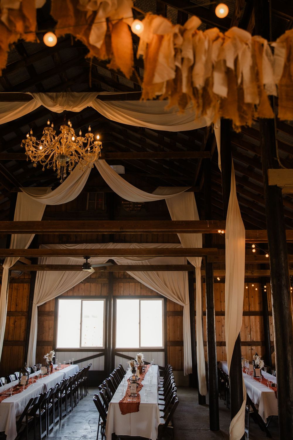 Rustic barn wedding reception venue with elegant white drapes, chandeliers, and long dining tables set for a celebration.