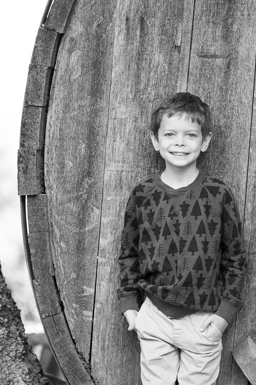Black and white portrait of a child wearing a patterned sweater standing against a rustic wooden barrel near San Jose, California