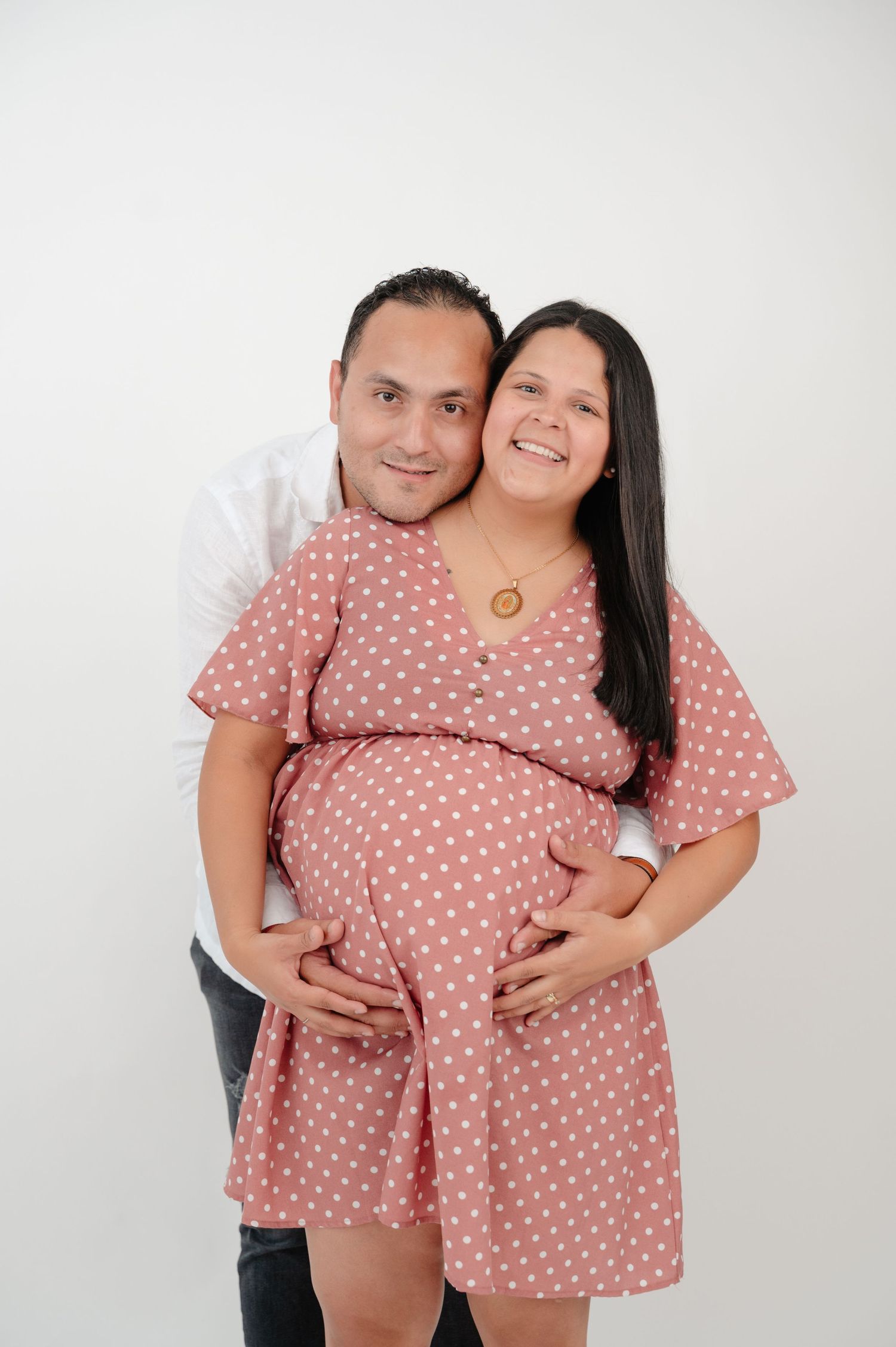 A couple wearing matching pink polka dot outfits pose together in a maternity photoshoot against a white backdrop.