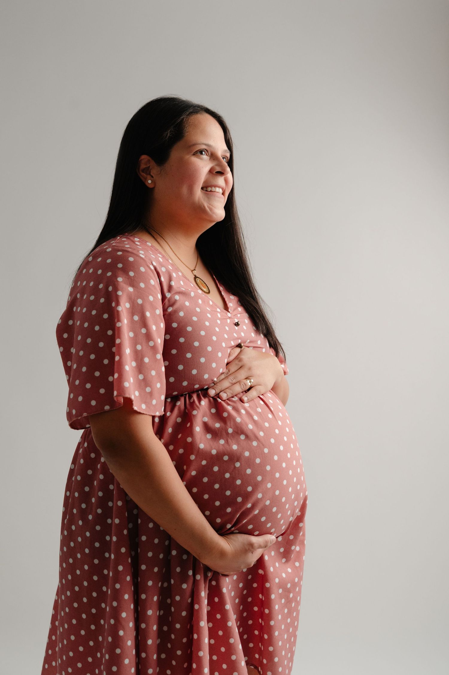 A pregnant woman in a pink polka dot dress poses for maternity photos against a light background.