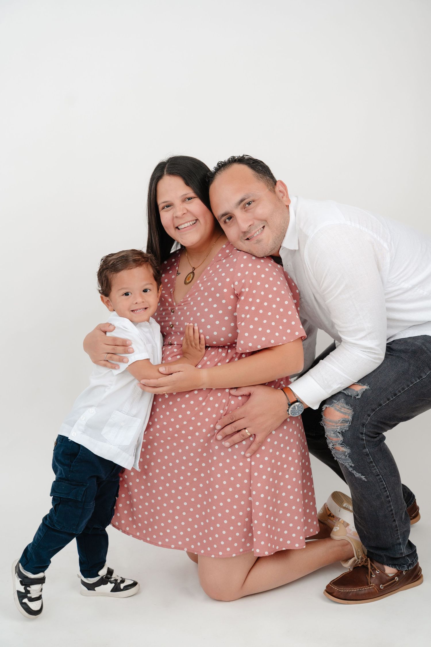 A family poses together against a white backdrop, with a young child in white and a woman wearing a pink polka dot dress.