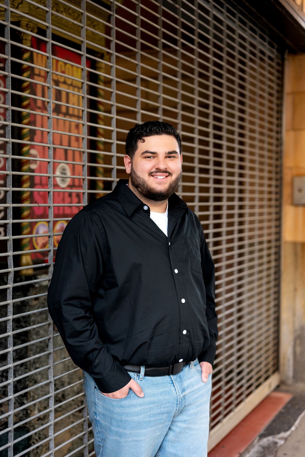 Smiling young man in black shirt leaning against a metal security gate on a city street.