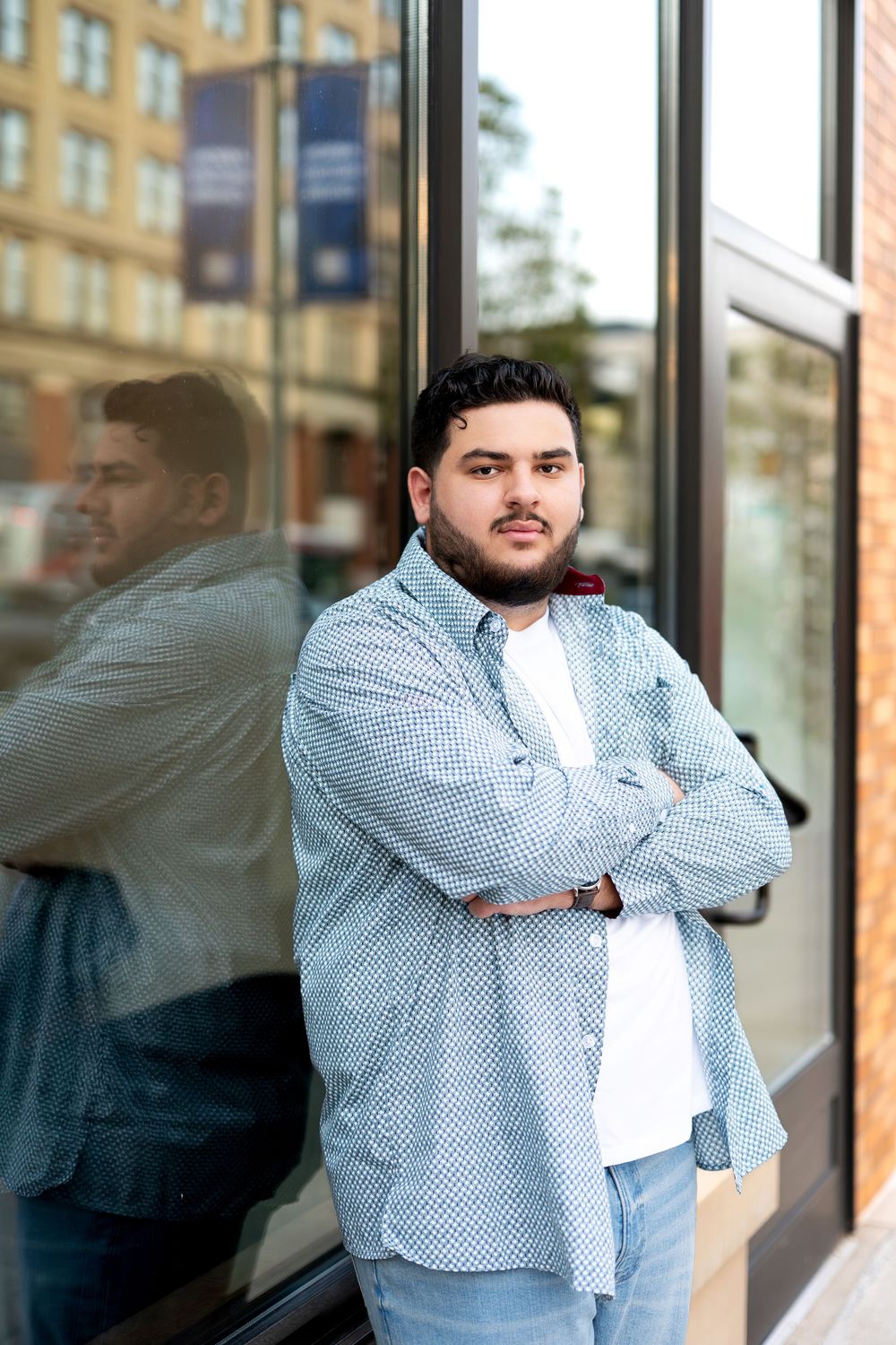 Man in blue patterned shirt standing with arms crossed against a glass building exterior.