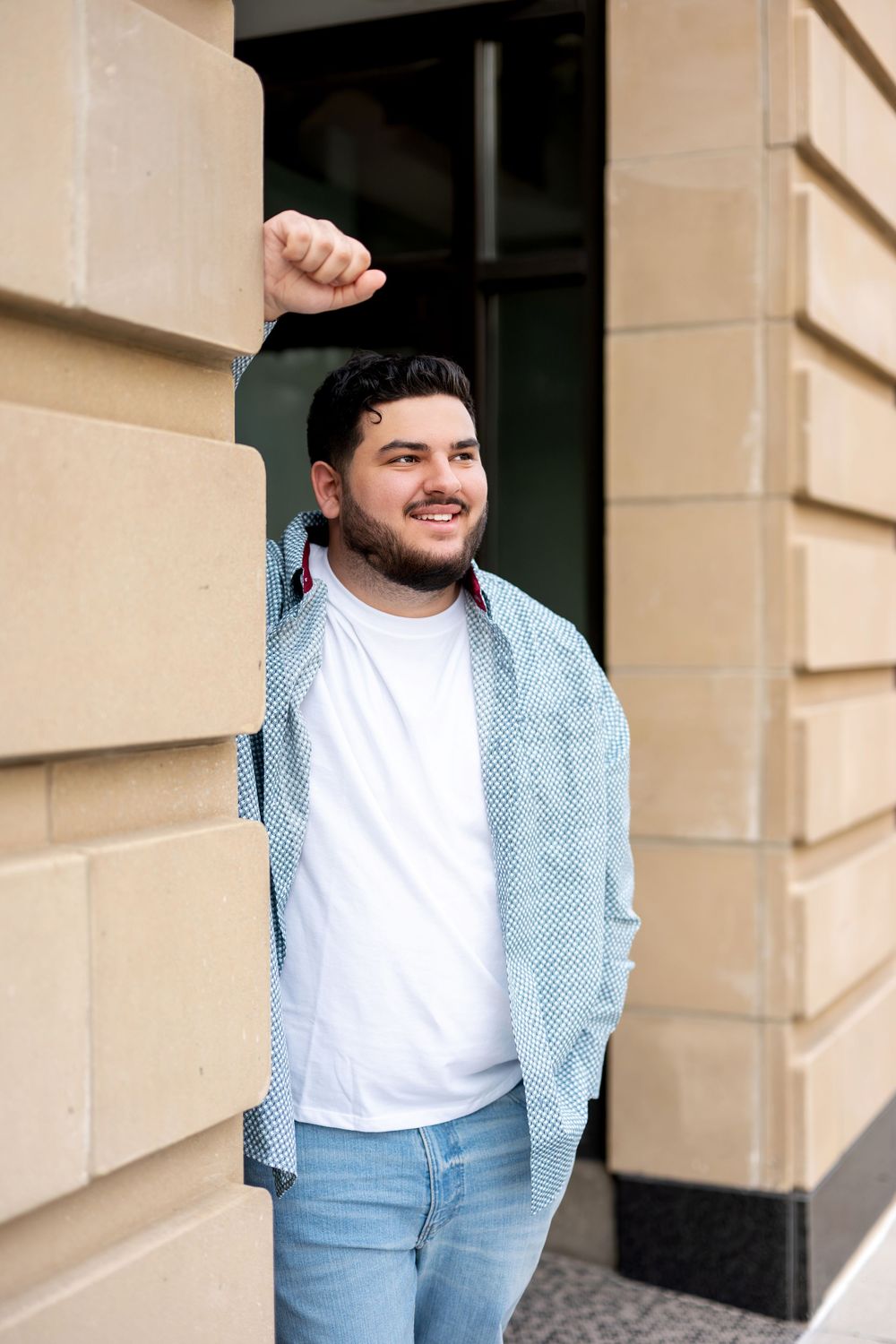 Smiling young man with beard leaning against stone building pillar wearing blue patterned shirt and white tee.