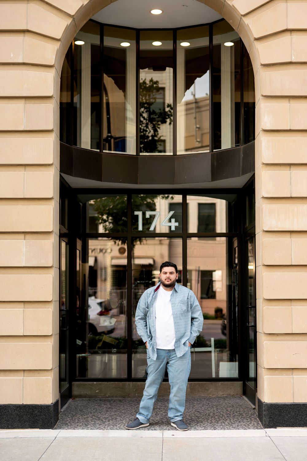 Young bearded man standing confidently in front of building entrance numbered 174 in urban setting.