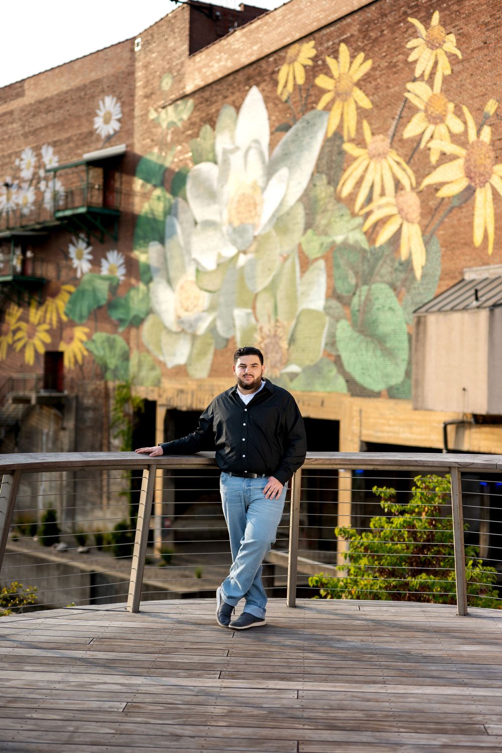 Man in black varsity jacket posing on a wooden walkway with a colorful flower mural backdrop.