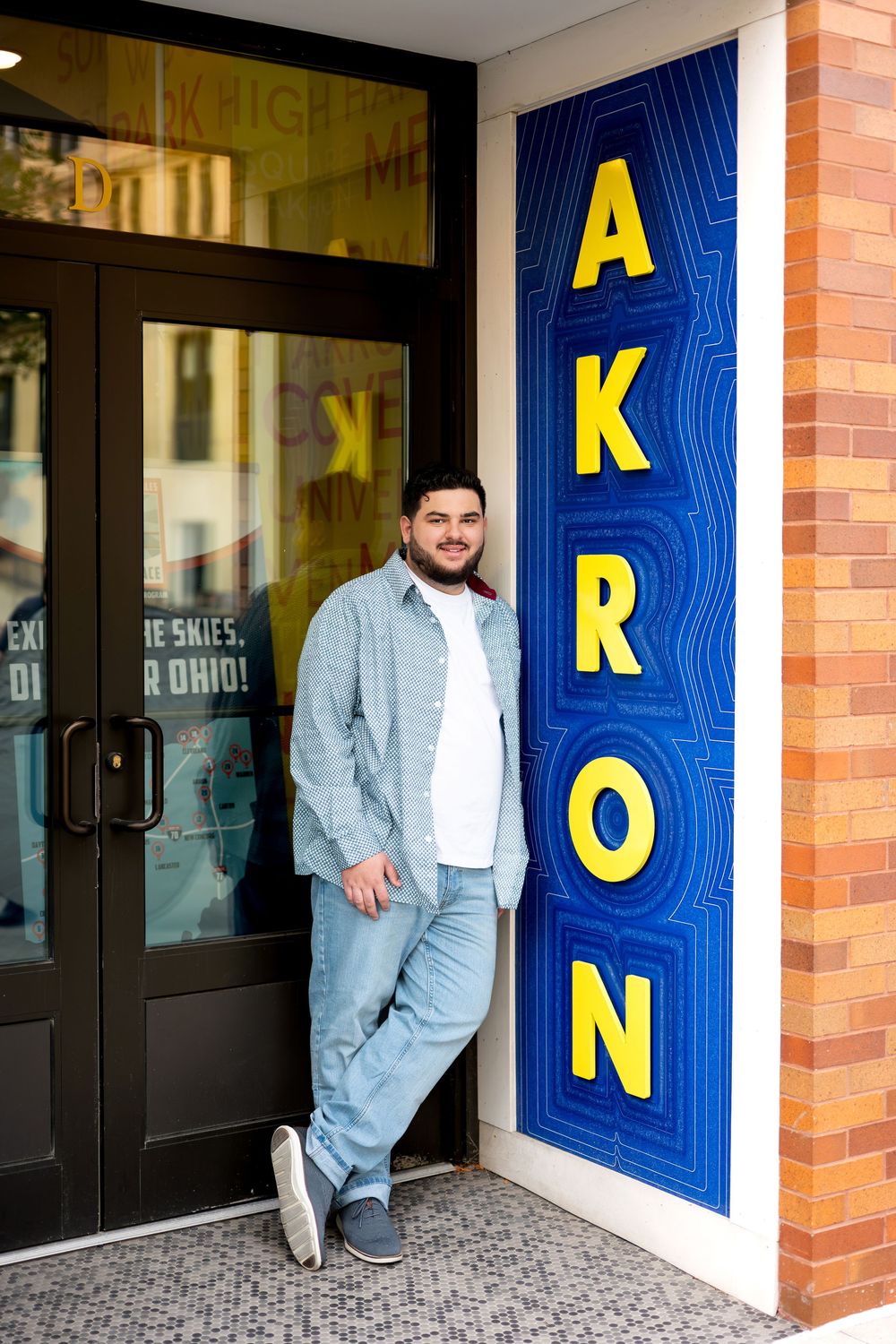 Smiling young man leaning against colorful Akron sign outside building entrance in casual outfit.