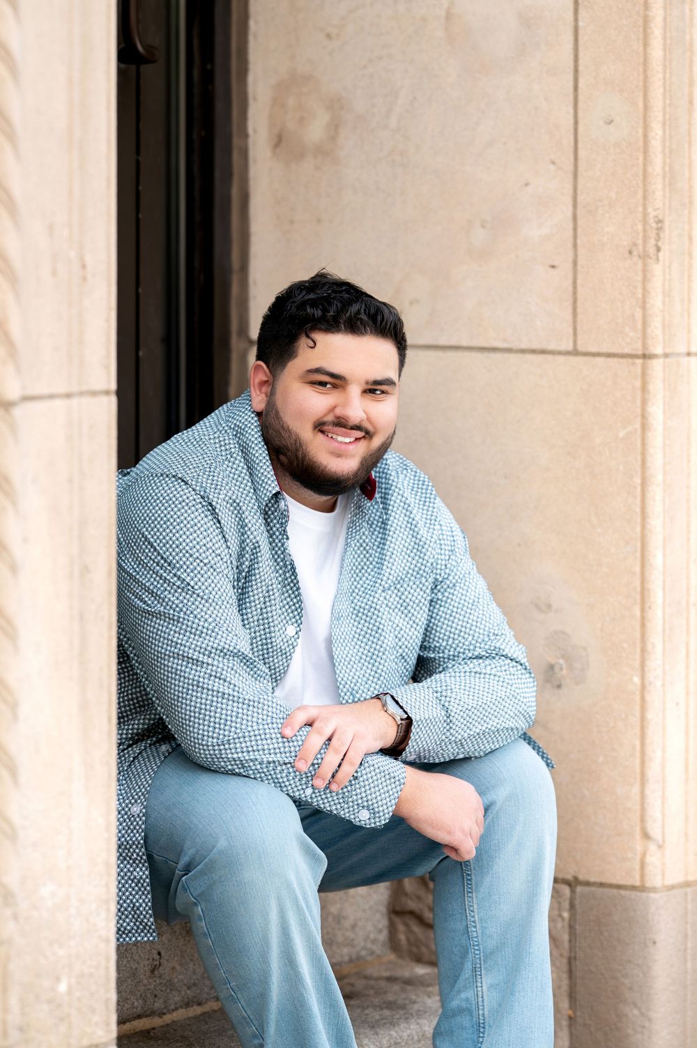 Smiling young bearded man sitting relaxed on stone steps wearing blue patterned shirt and jeans.