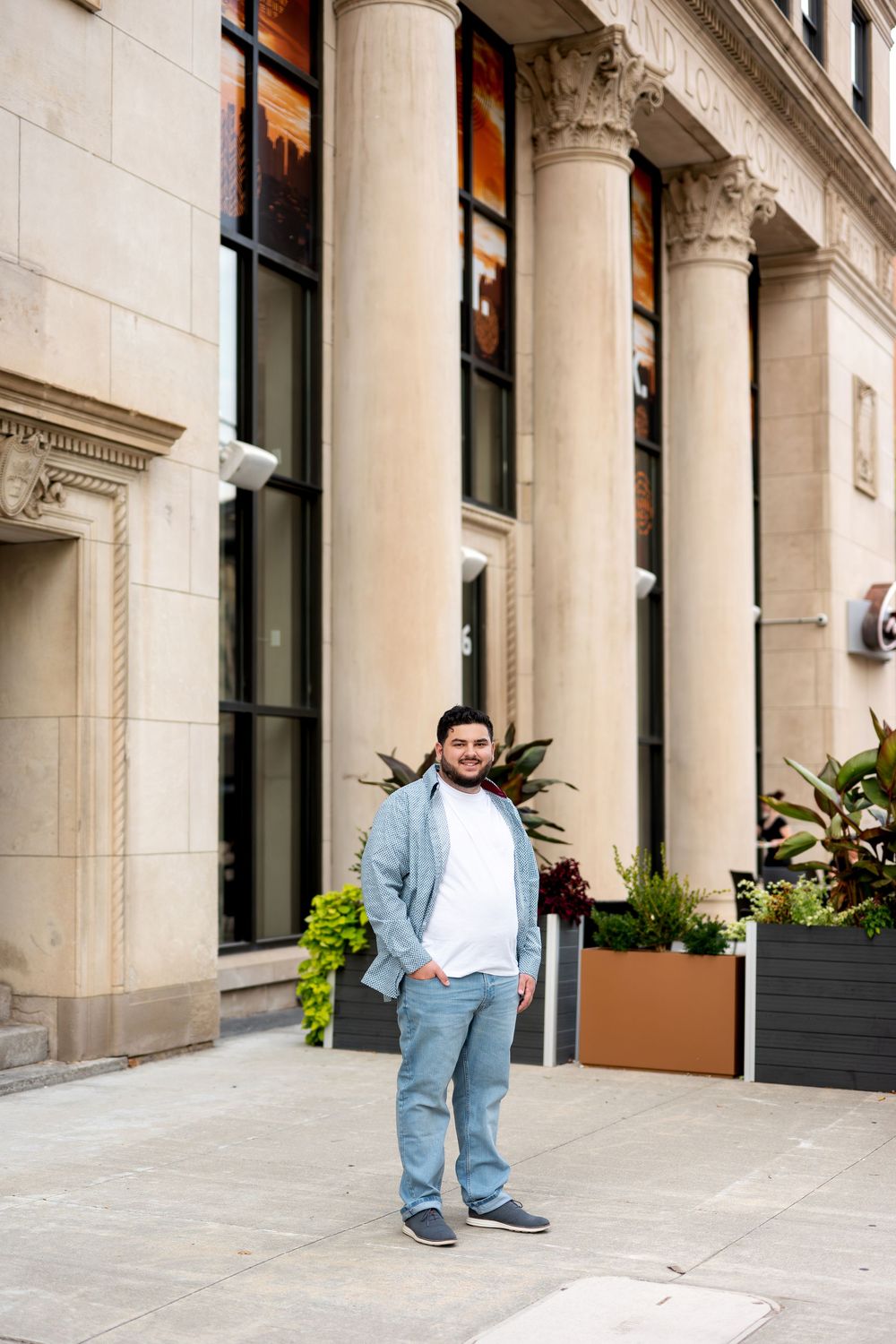 Smiling young man in blue shirt and jeans posing on sidewalk beside classical columned building.