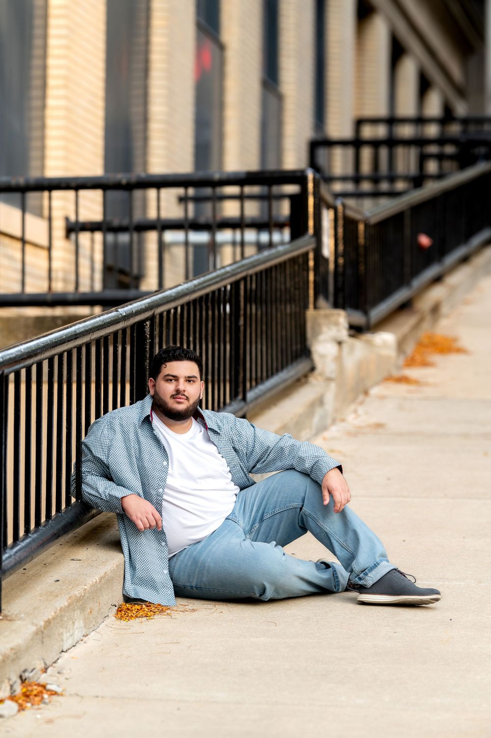 Young man in light blue outfit sitting relaxed against urban railing on city sidewalk.