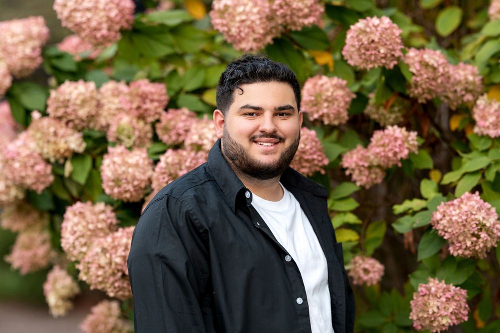 Smiling young man in black jacket posing in front of blooming pink hydrangea bushes outdoors.
