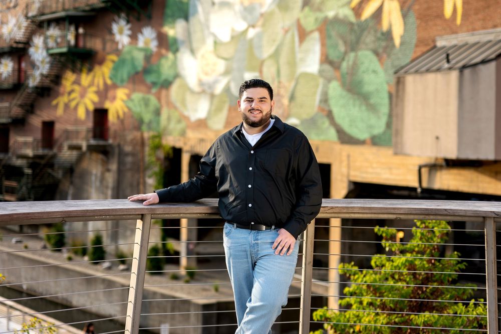 Smiling young man in black shirt leaning on a metal railing with a colorful floral mural behind.