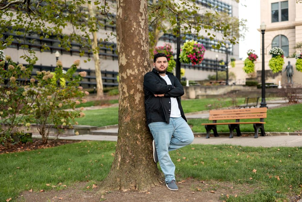 Young man in black jacket leaning against large tree in urban park with bench in background.