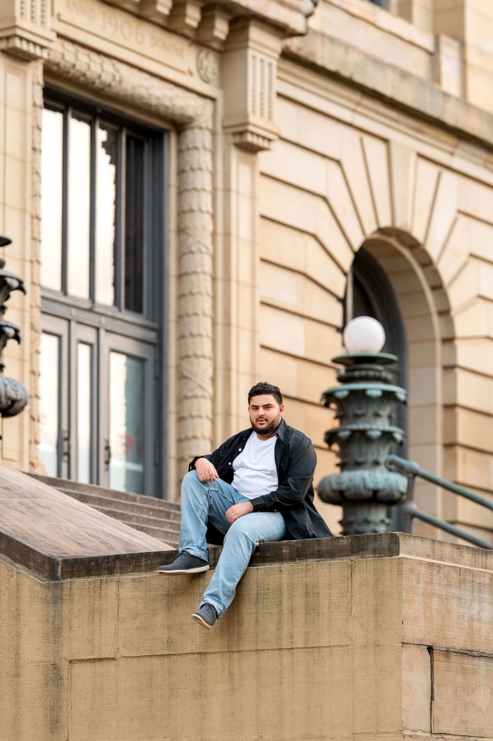 Young man in black jacket and jeans sits relaxed on stone ledge of classic building.