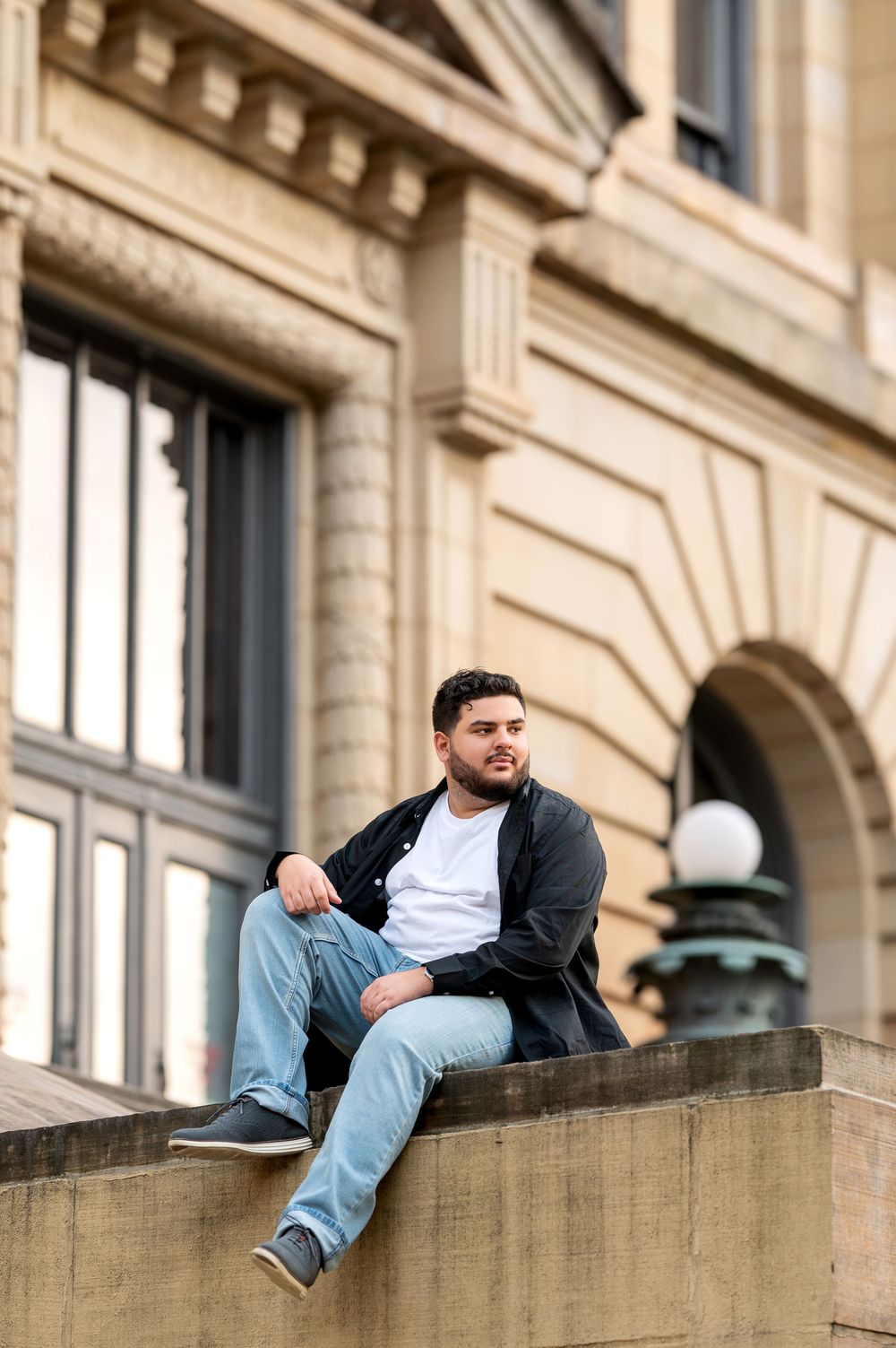 Young man in black jacket sits on stone ledge of grand historic beige building exterior.