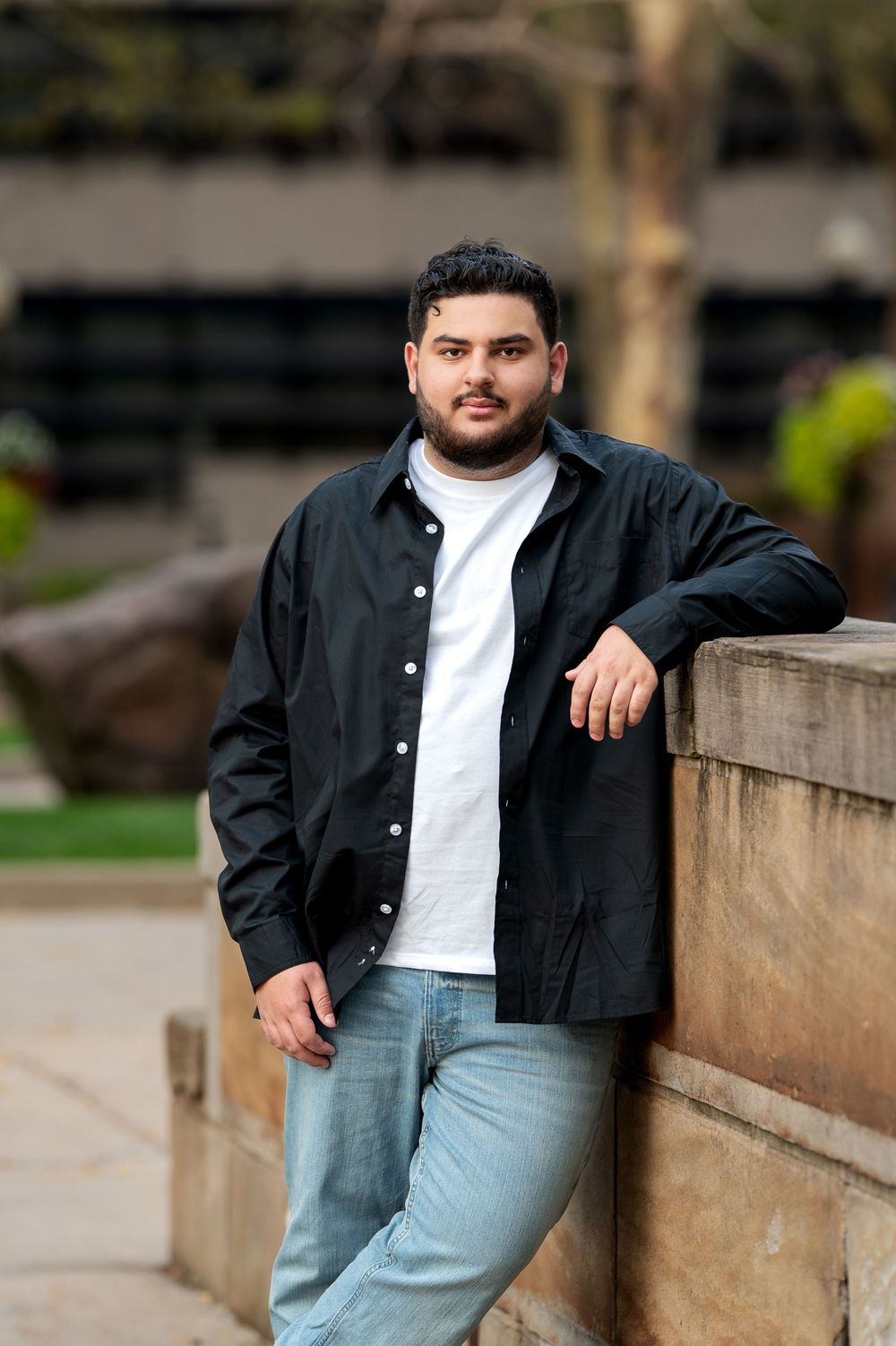 Young man in black shirt and jeans leaning against stone wall outdoors.