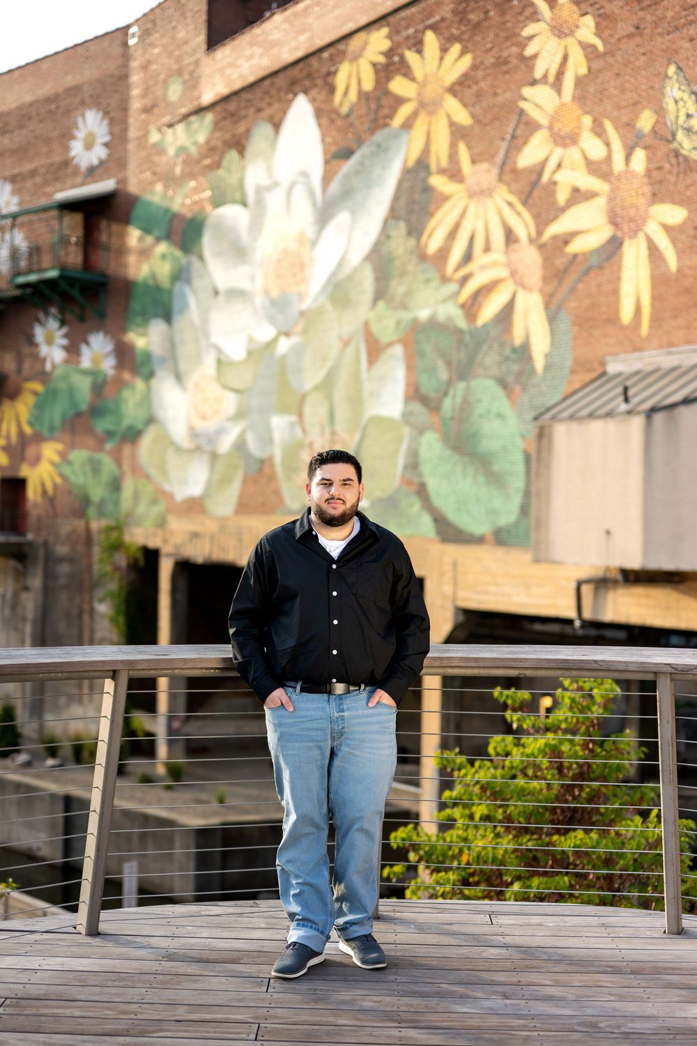 Man in black jacket and jeans standing on wooden bridge with large flower mural behind him.