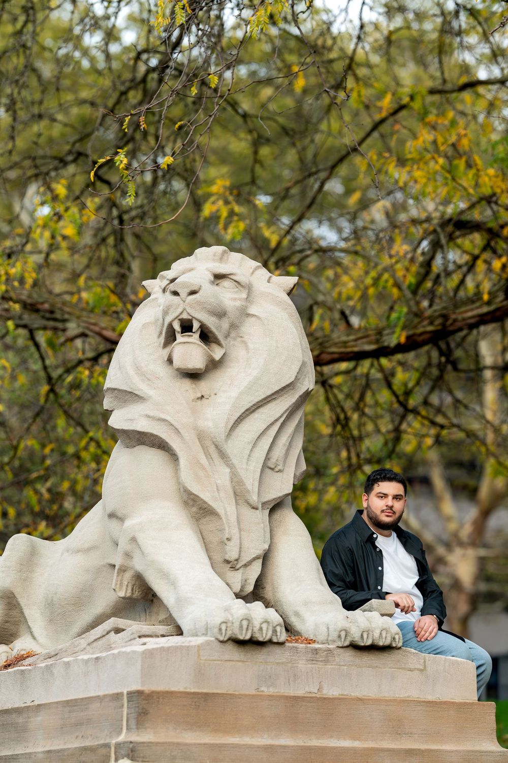 Man sitting beside a large stone lion statue with open mouth in a wooded park setting.