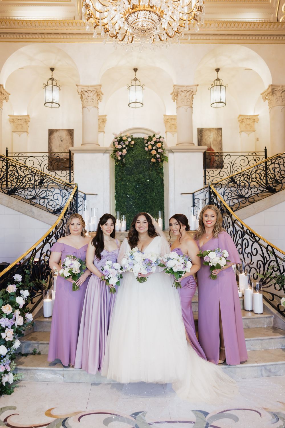 Wedding party in lavender dresses posing on an elegant staircase with ornate railings.