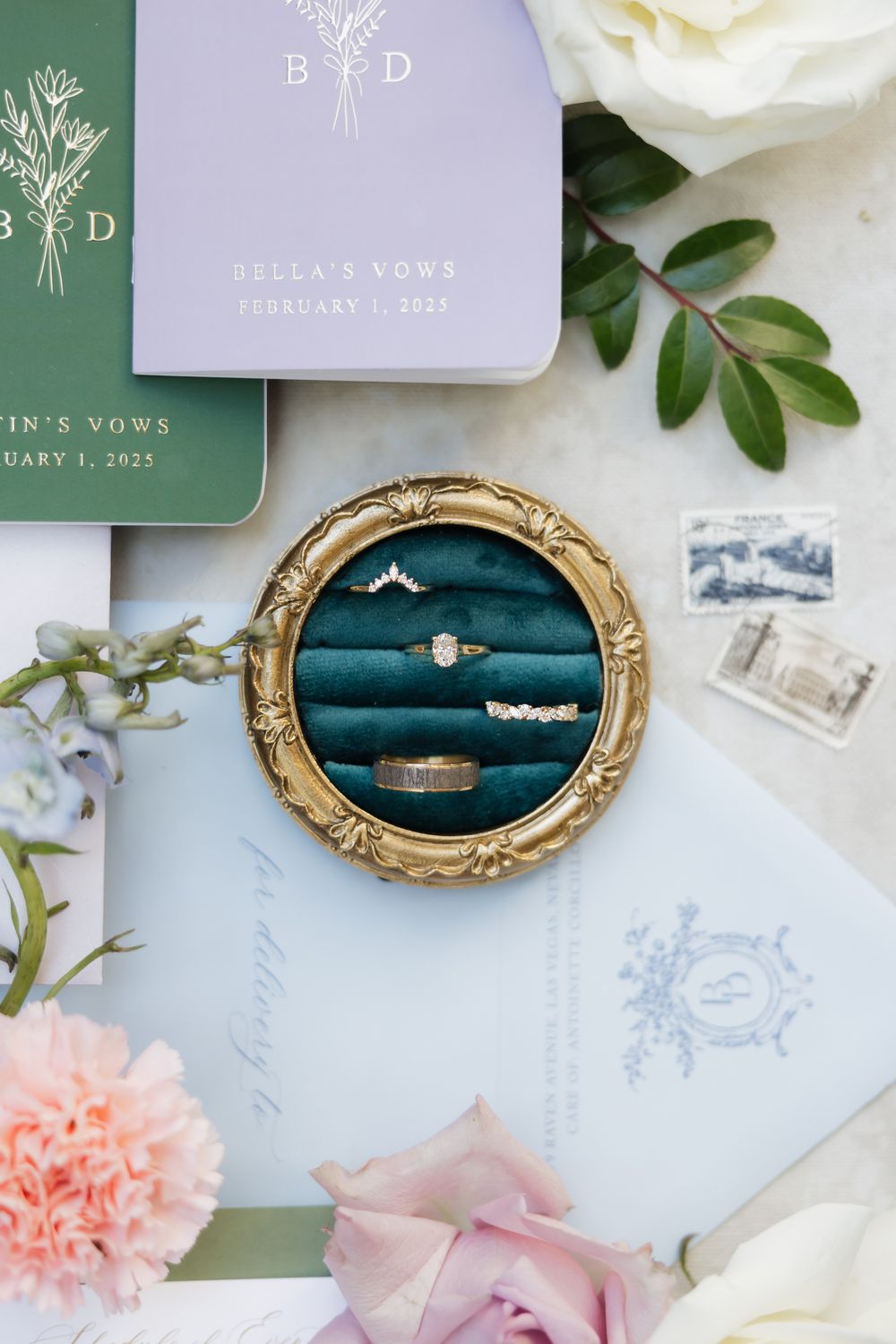 Wedding details featuring ornate gold frames with rings on teal velvet alongside purple and pink floral accents.
