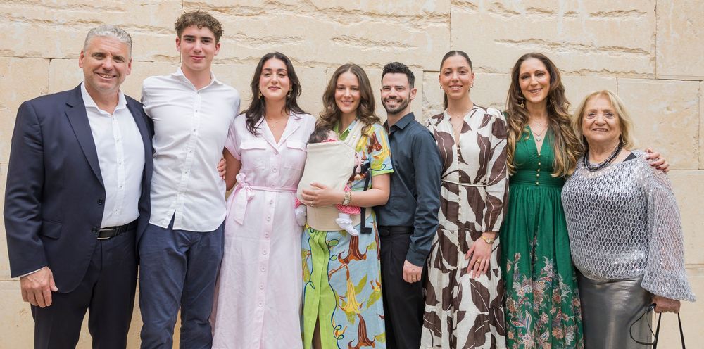 A large group of people in formal attire pose together for a celebratory photo against a light stone wall.
