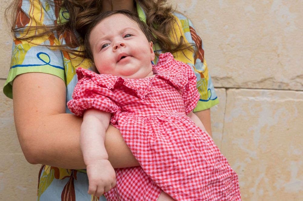 Baby wearing red gingham dress lies on beige floor while smiling and playing.