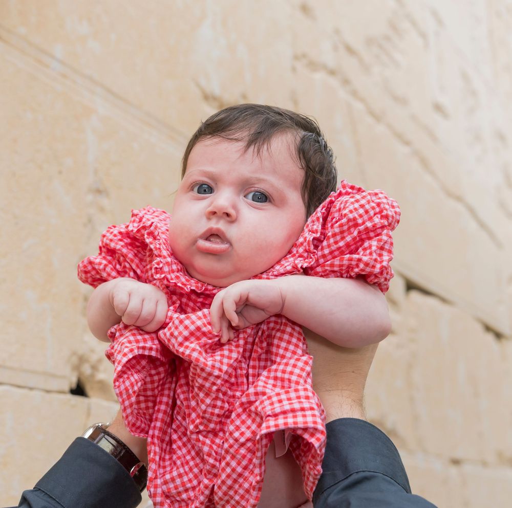 Baby wearing red and white gingham dress held up against beige wall background.
