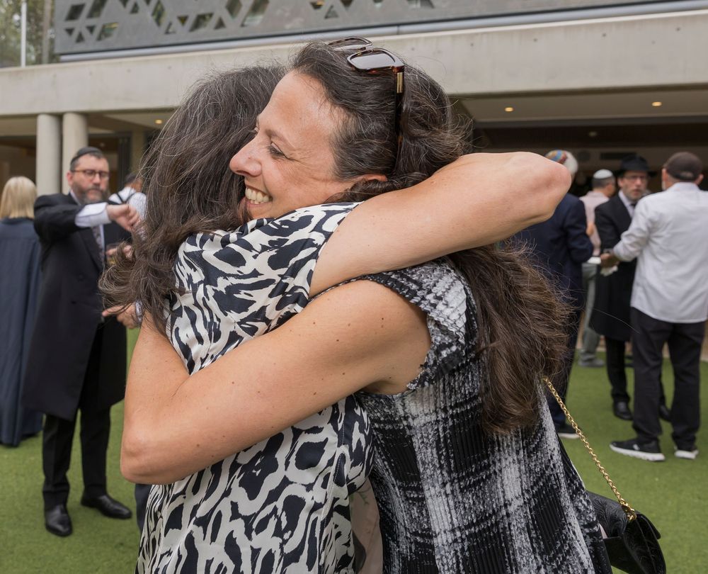 Two people embrace in a celebratory hug at an outdoor event with other attendees visible in the background.