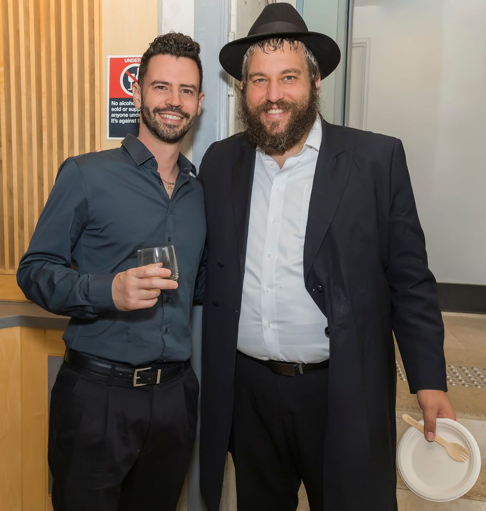 Two men in formal attire standing together at an event, one wearing a black hat and the other in a dark suit.
