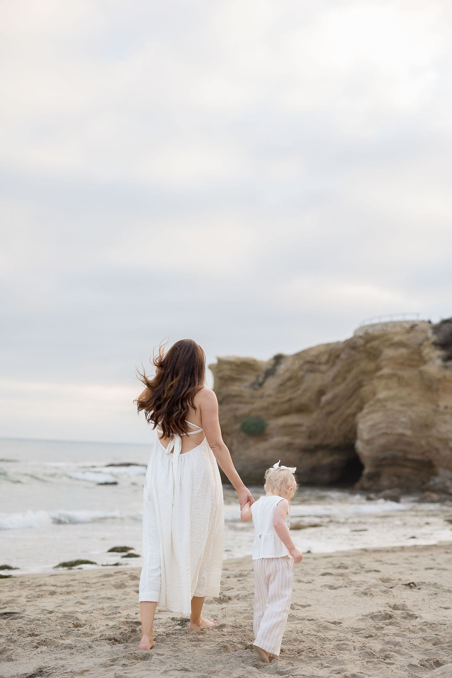 A mother and child in white dresses share a playful moment together while lying on a sandy beach.